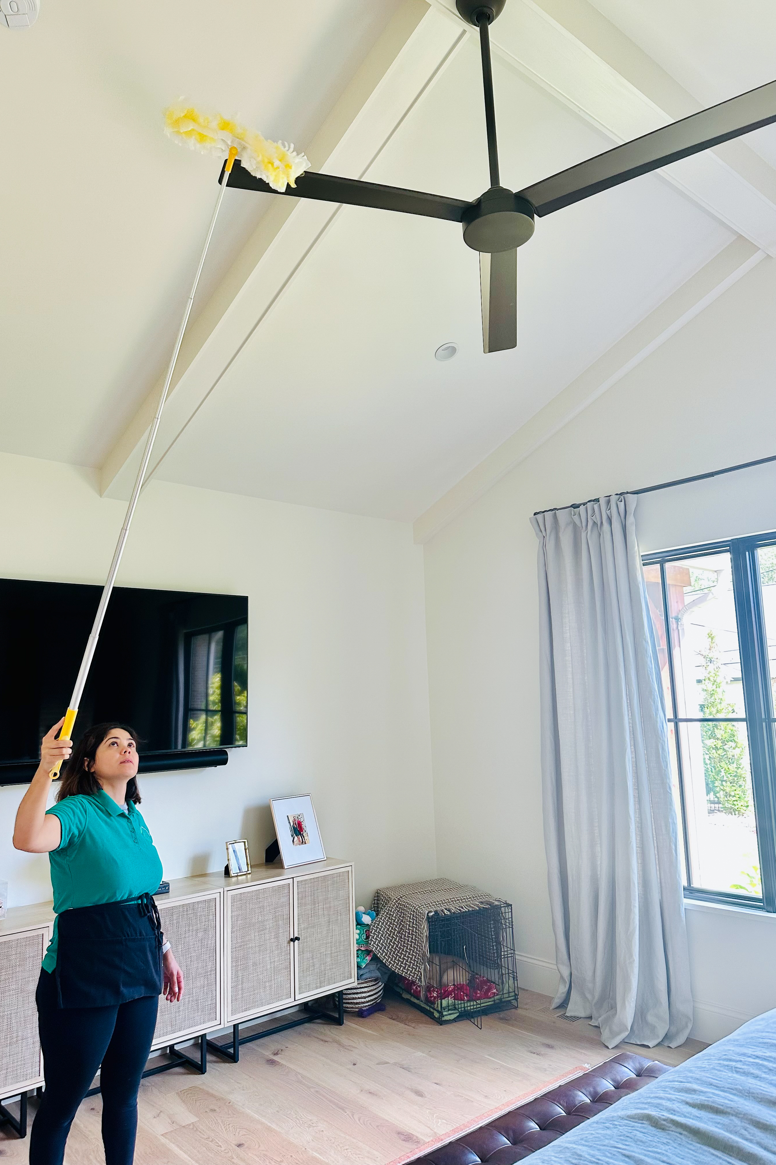 rofessional cleaner using an extended duster to clean a ceiling fan, showcasing meticulous attention to detail in a bright, well-organized room.