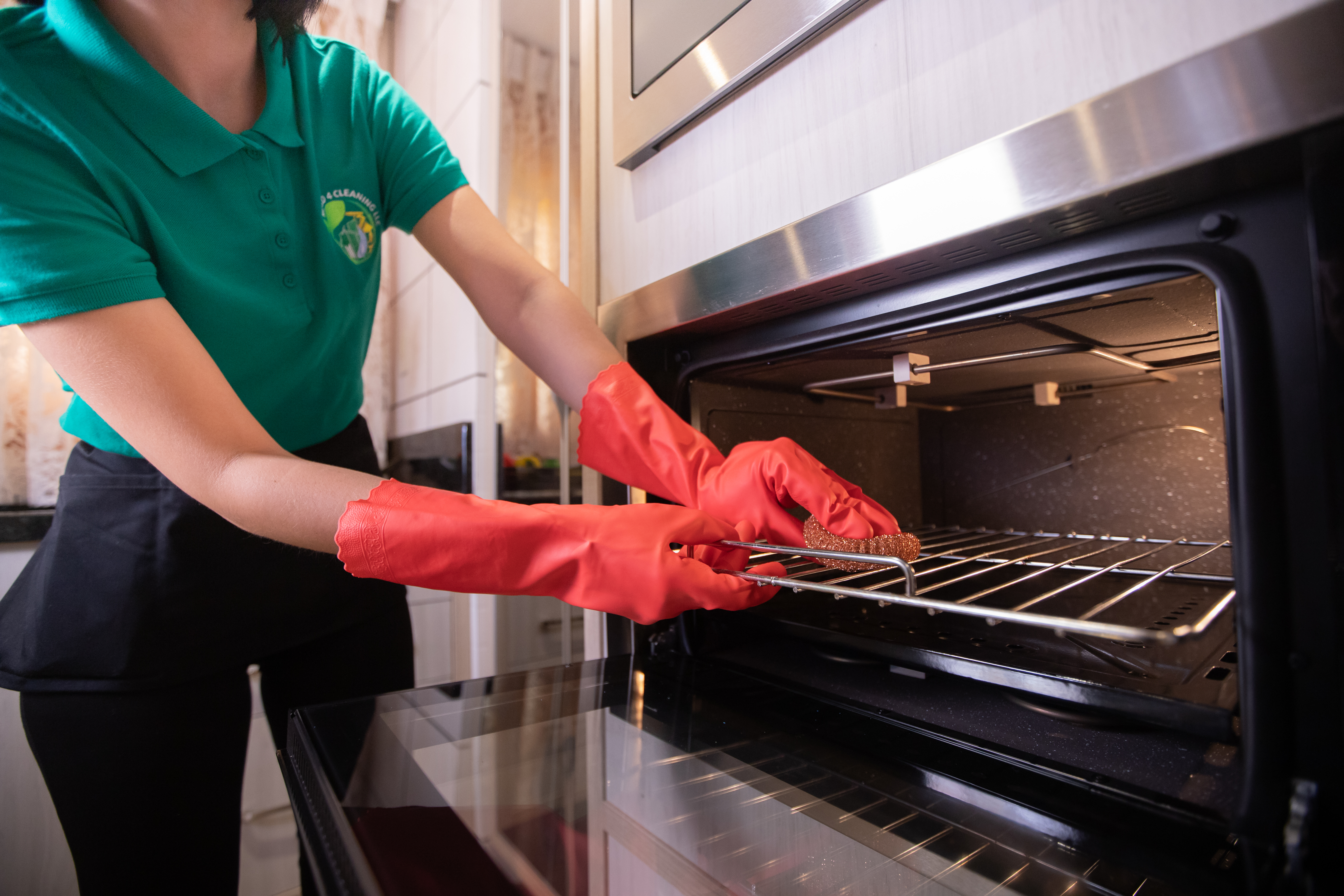 A Maid 4 Cleaning professional wearing a branded green uniform and red gloves carefully scrubs an oven rack, demonstrating meticulous attention to detail and thorough kitchen cleaning services.