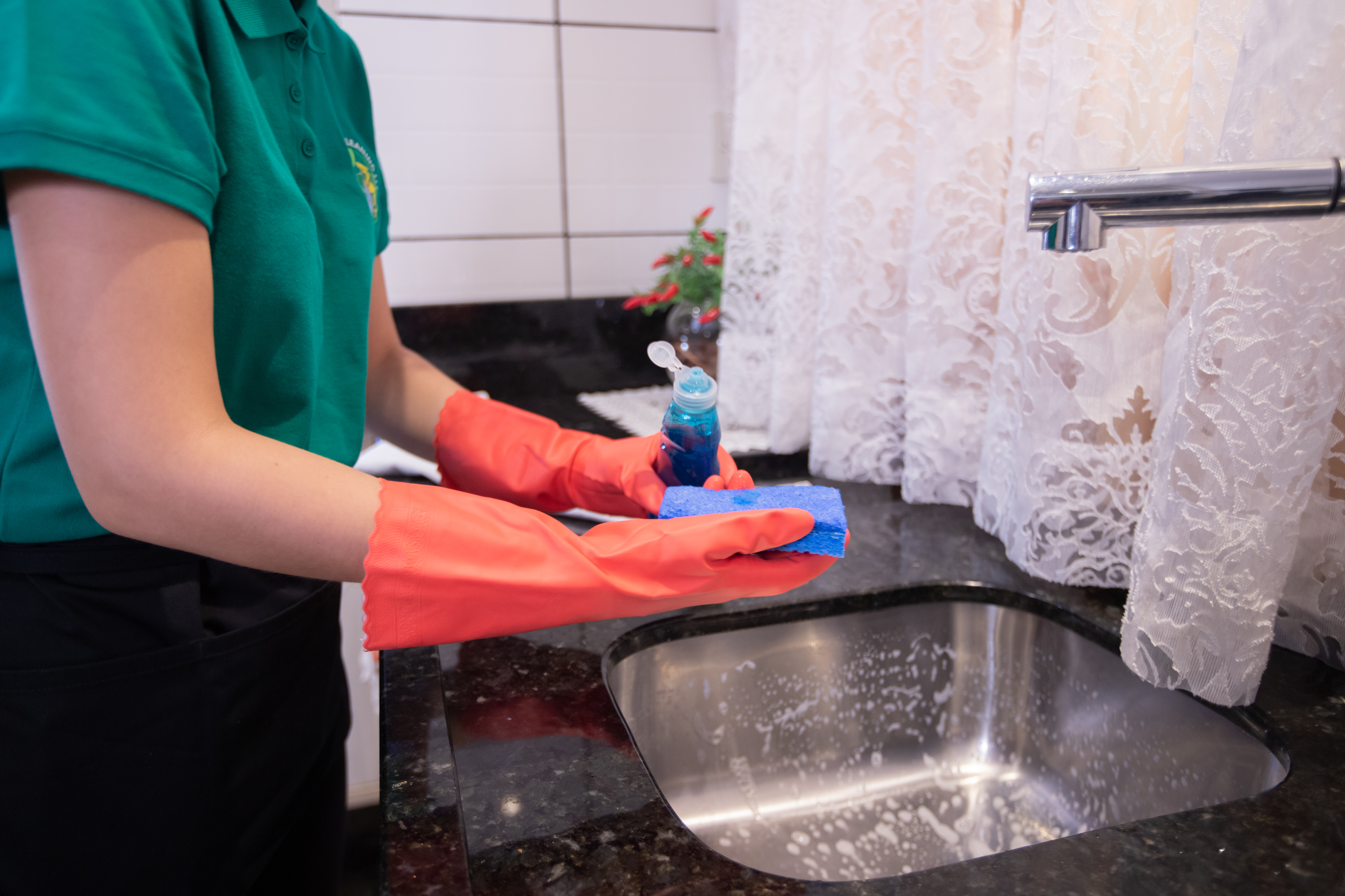 A Maid 4 Cleaning professional wearing orange gloves applies dish soap onto a sponge, preparing to thoroughly clean a kitchen sink, showcasing the company's dedication to detailed and effective home cleaning.