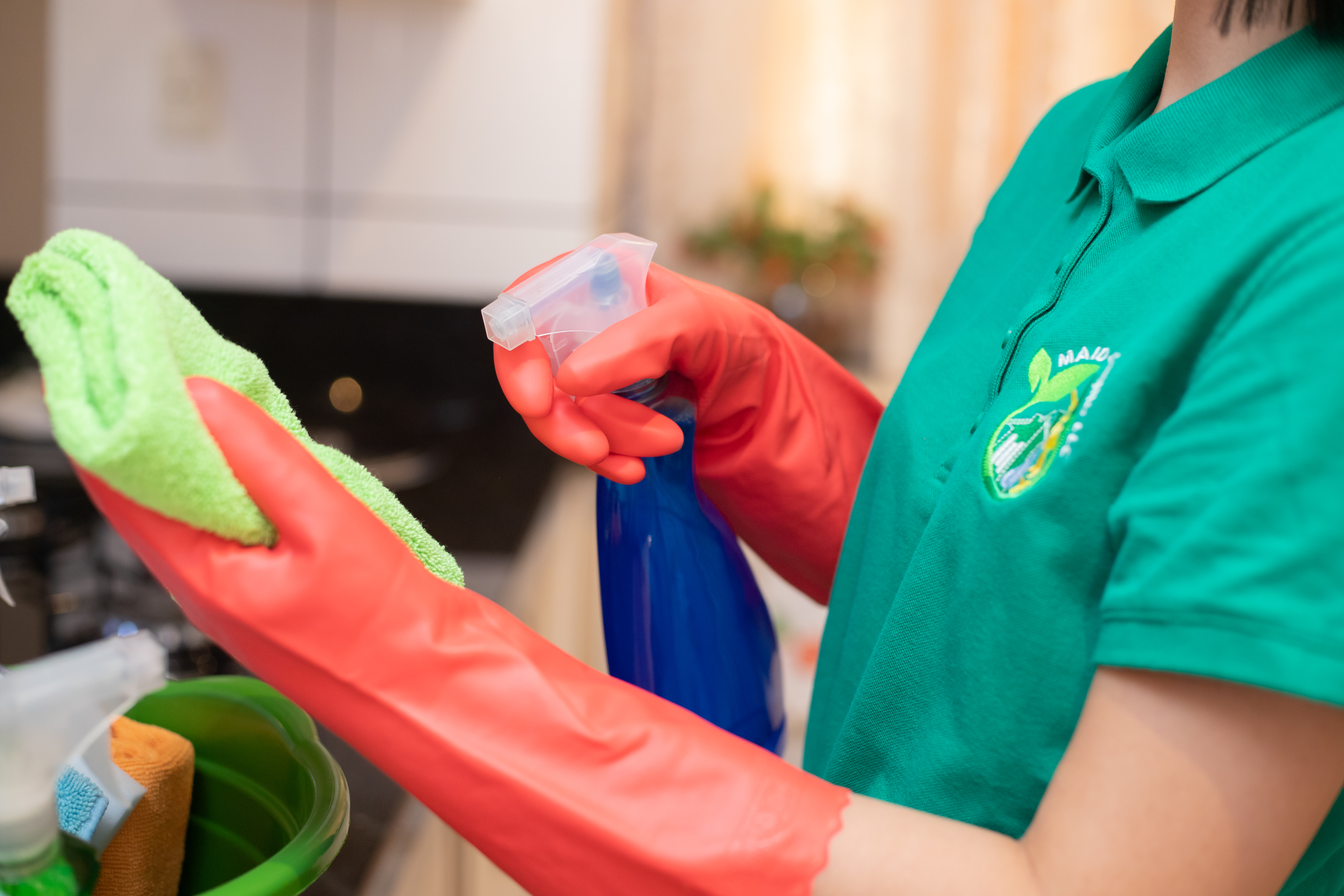 A Maid 4 Cleaning professional wearing red gloves and a branded green polo sprays cleaner onto a green microfiber cloth, ready to clean a kitchen microwave