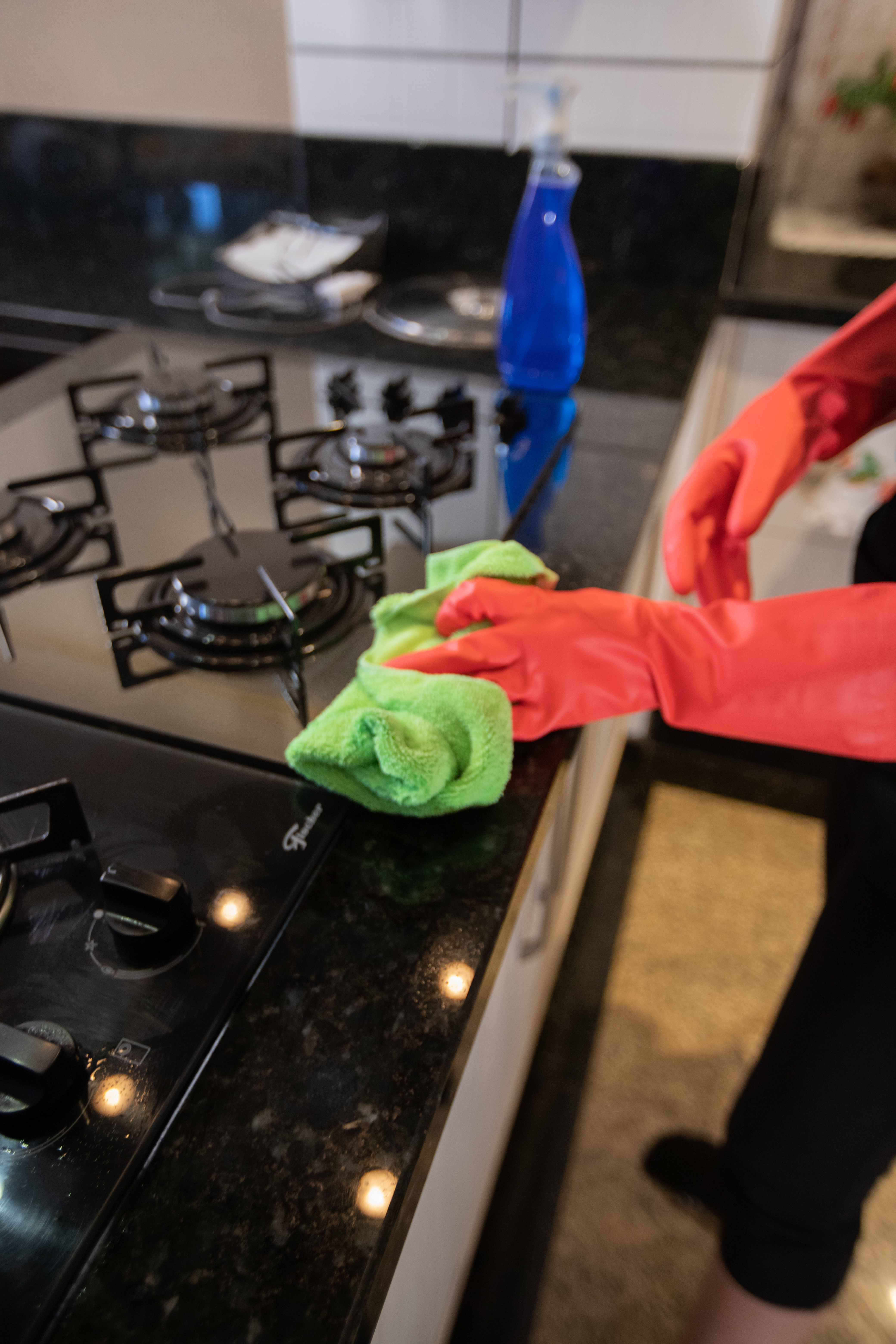 A person wearing red rubber gloves cleaning a black granite kitchen countertop and stovetop with a green microfiber cloth. A spray bottle with a blue cleaning solution is visible in the background.