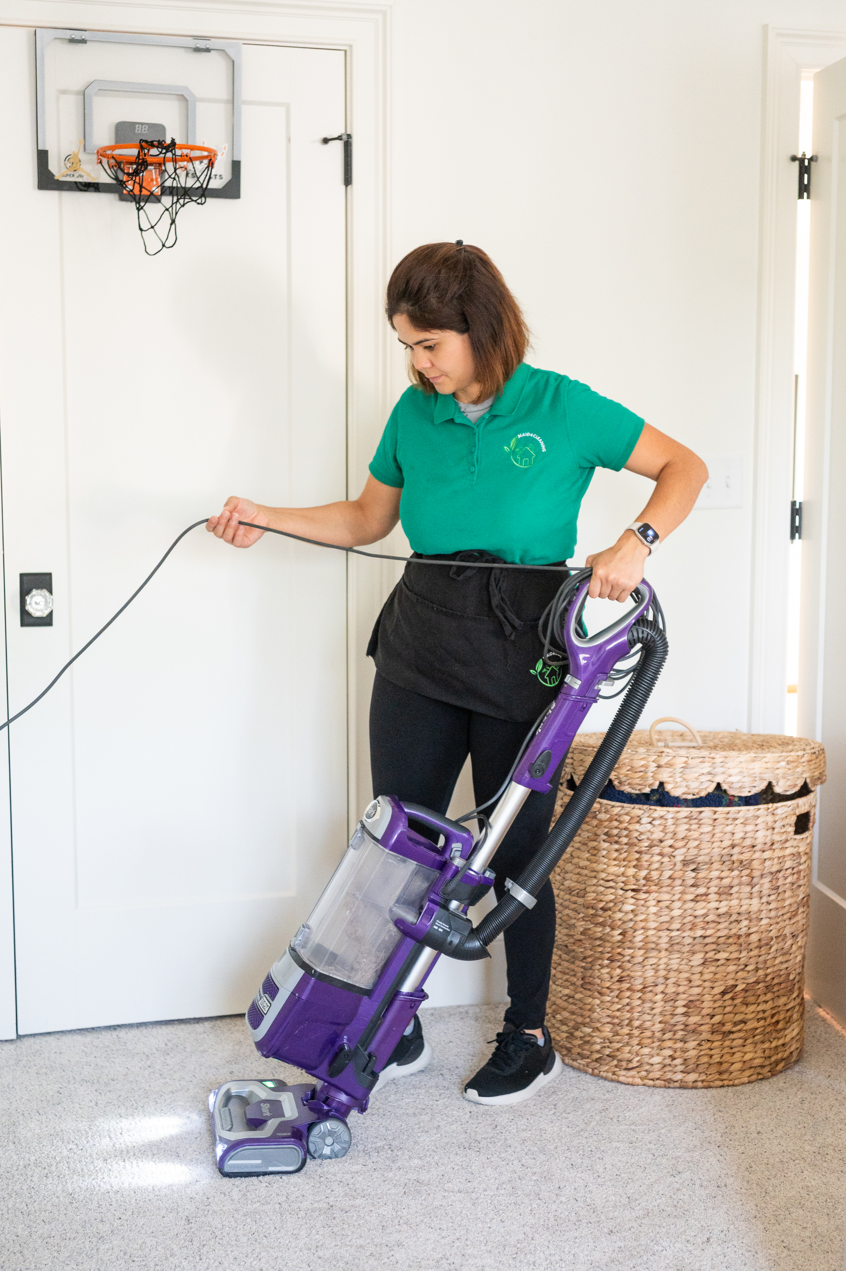 A professional cleaner wearing a green uniform shirt and black apron is vacuuming a beige carpet in a well-lit room. She is holding a purple upright vacuum cleaner while managing its power cord. A woven laundry basket is placed nearby, and a small basketball hoop hangs on a white door in the background.