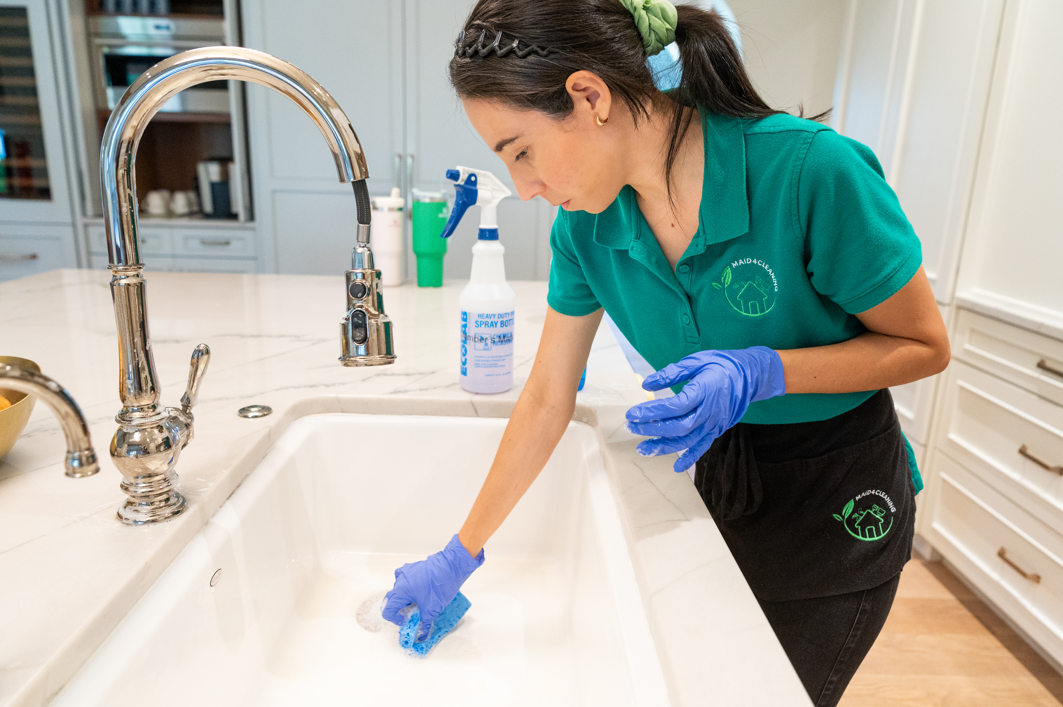 A professional cleaner wearing gloves and a green uniform is scrubbing a kitchen sink with a sponge, ensuring a sanitary and bacteria-free cleaning process. Cleaning supplies, including a disinfectant spray bottle, are visible in the background, emphasizing proper kitchen hygiene.
