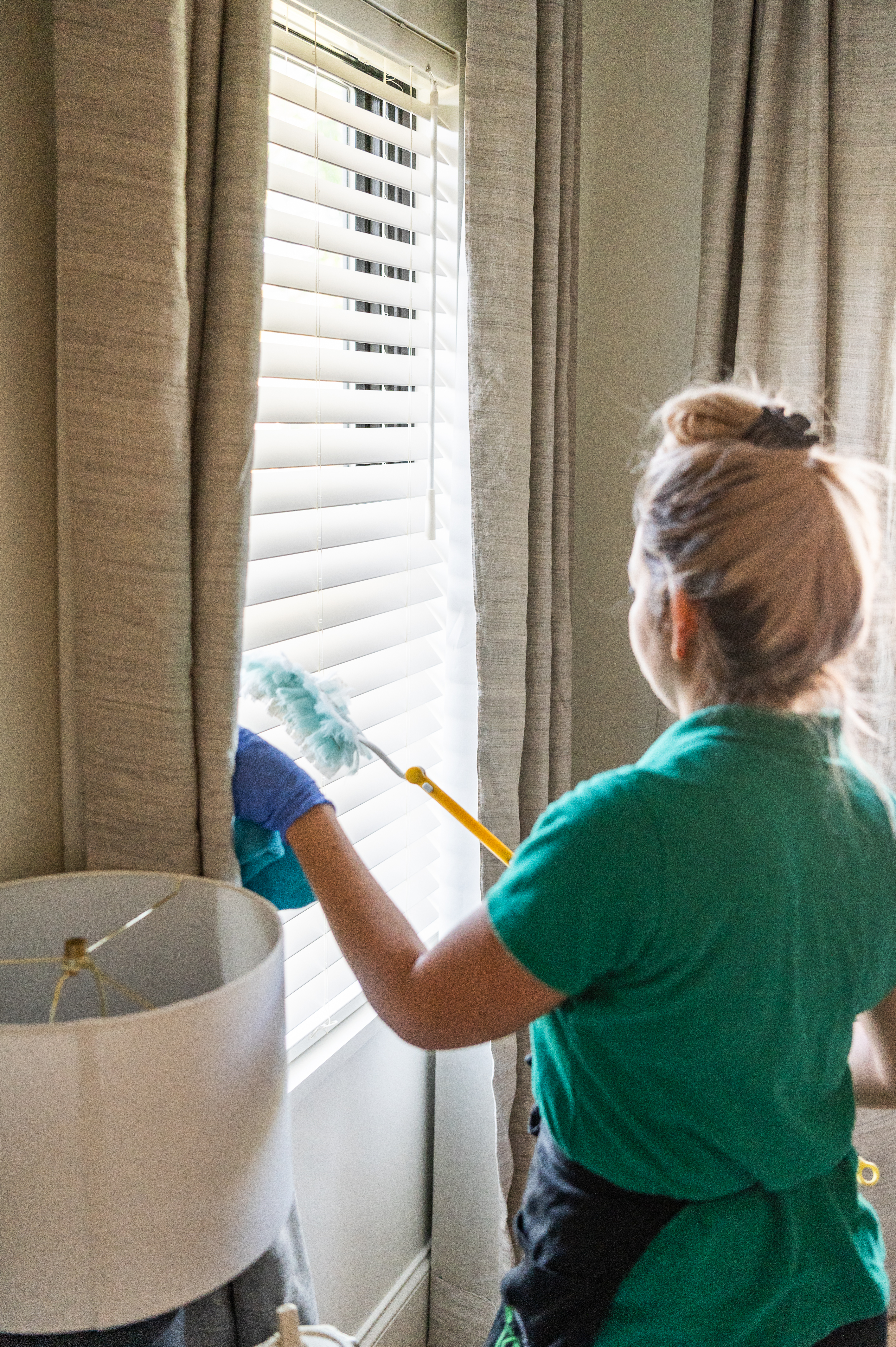 A professional cleaner wearing a green uniform and gloves is dusting white window blinds near beige curtains using a duster. A lamp is partially visible in the foreground. Natural light is filtering through the blinds, highlighting the clean and fresh space.