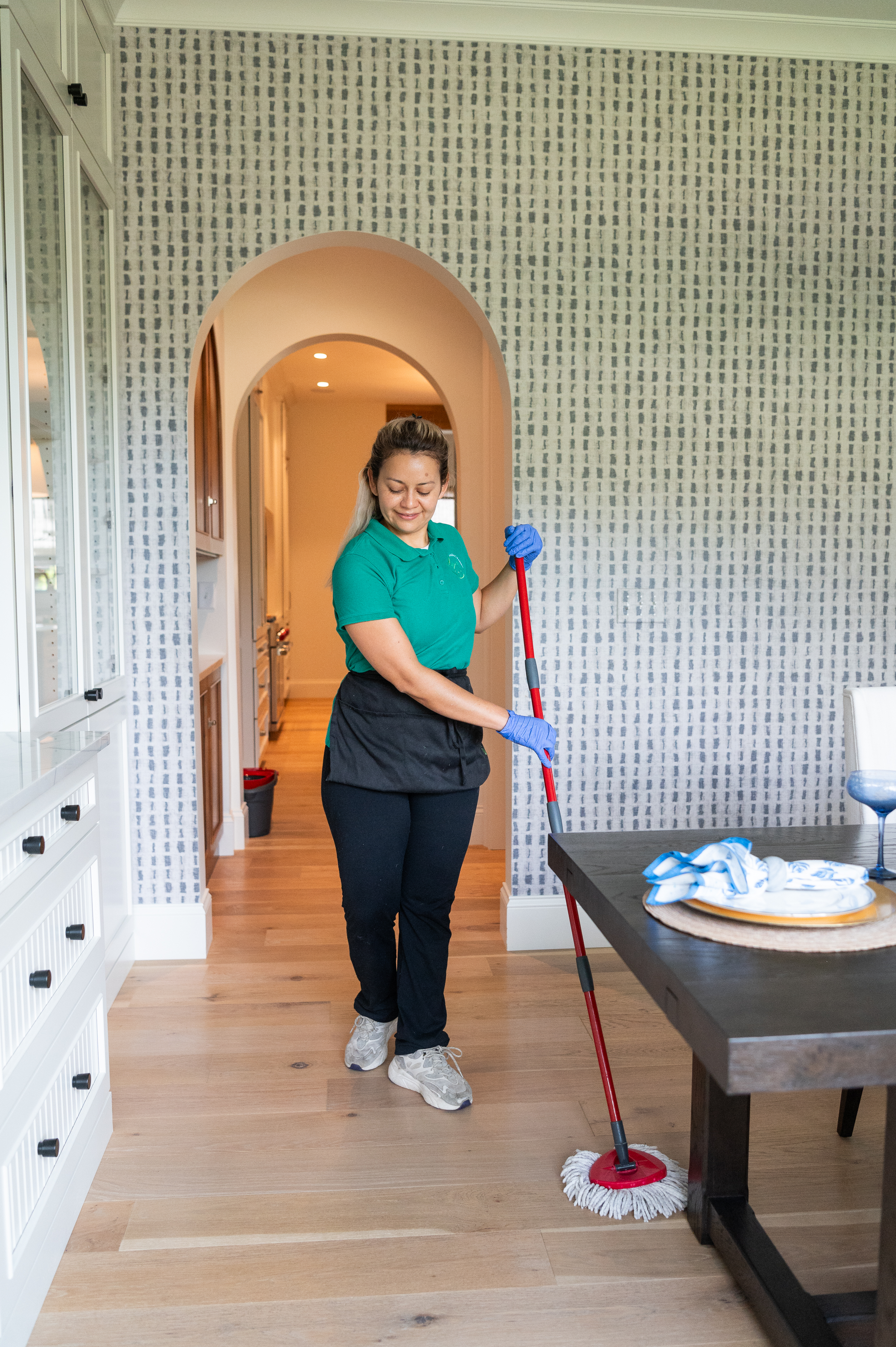 A professional cleaner wearing a green polo shirt, black apron, and blue gloves is mopping a hardwood floor in a well-lit, modern dining area. The room features a dark wooden dining table set with plates and napkins, white cabinetry, and a stylish arched entryway leading to a kitchen in the background.