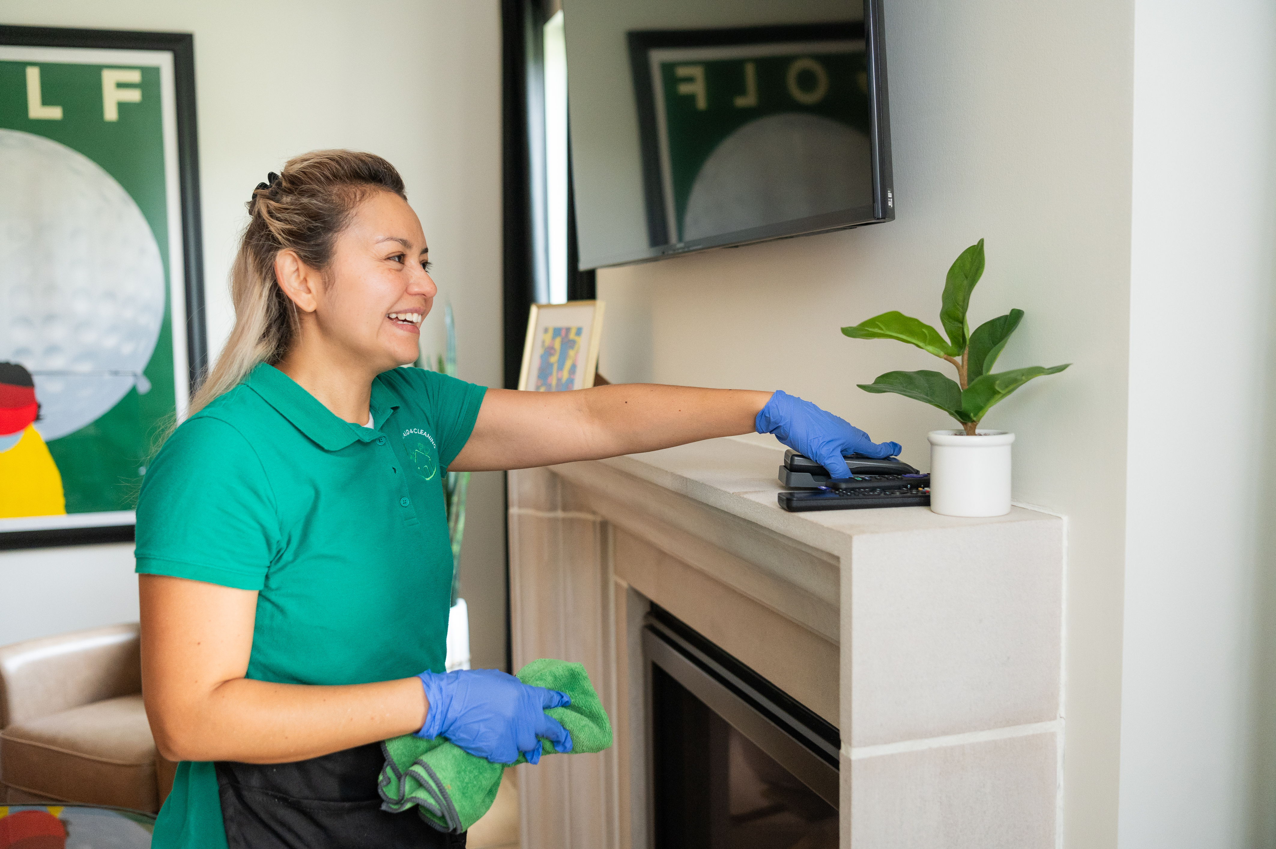 A professional cleaner wearing a green uniform and blue gloves dusting a fireplace mantel with a green microfiber cloth, ensuring a tidy and fresh living space. A small potted plant and remote controls are neatly arranged on the mantel.