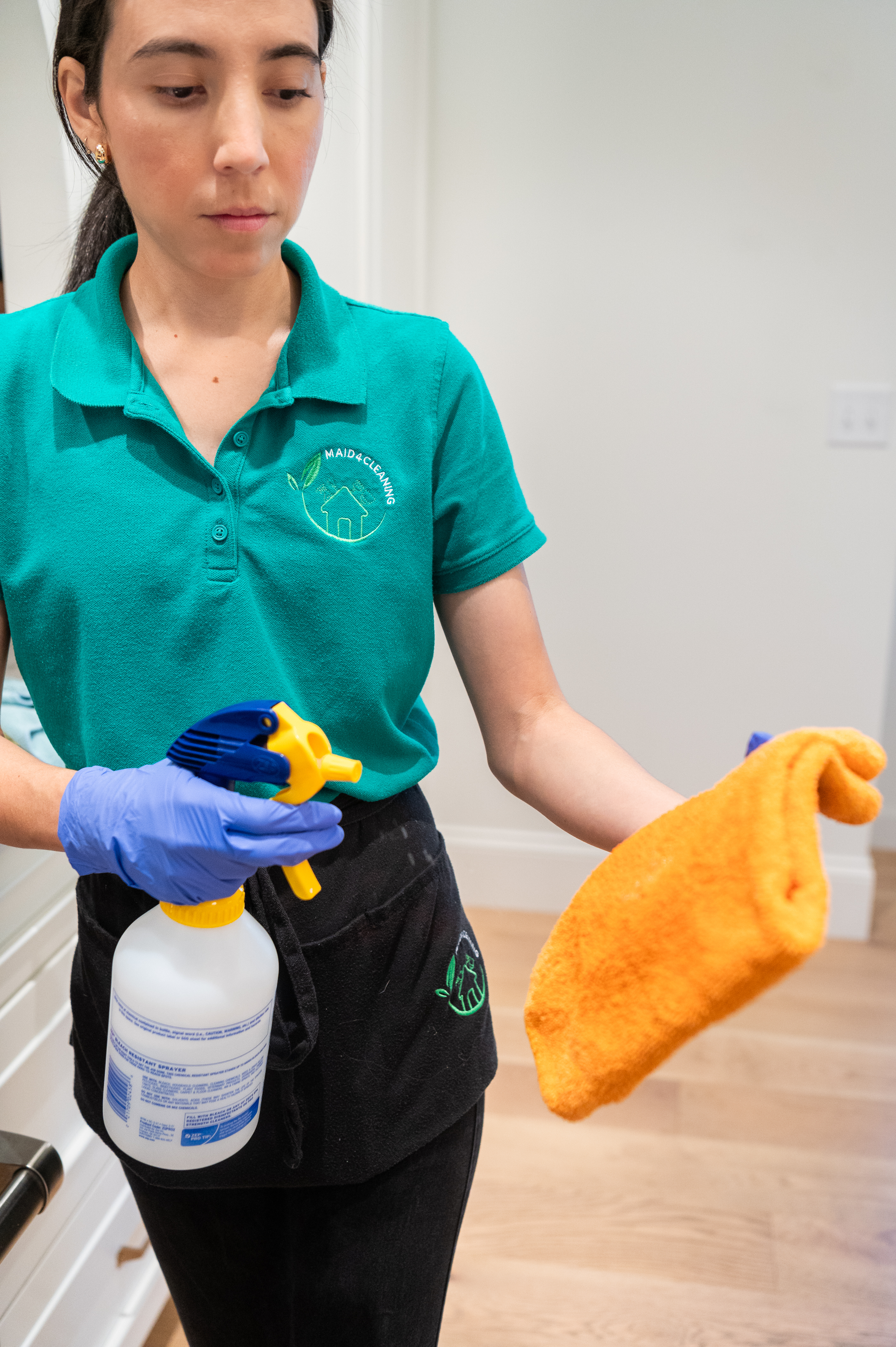 A professional maid wearing gloves and a green uniform uses a spray bottle and sponge to clean a home, showcasing the use of DIY cleaning products for a fresh and chemical-free environment.