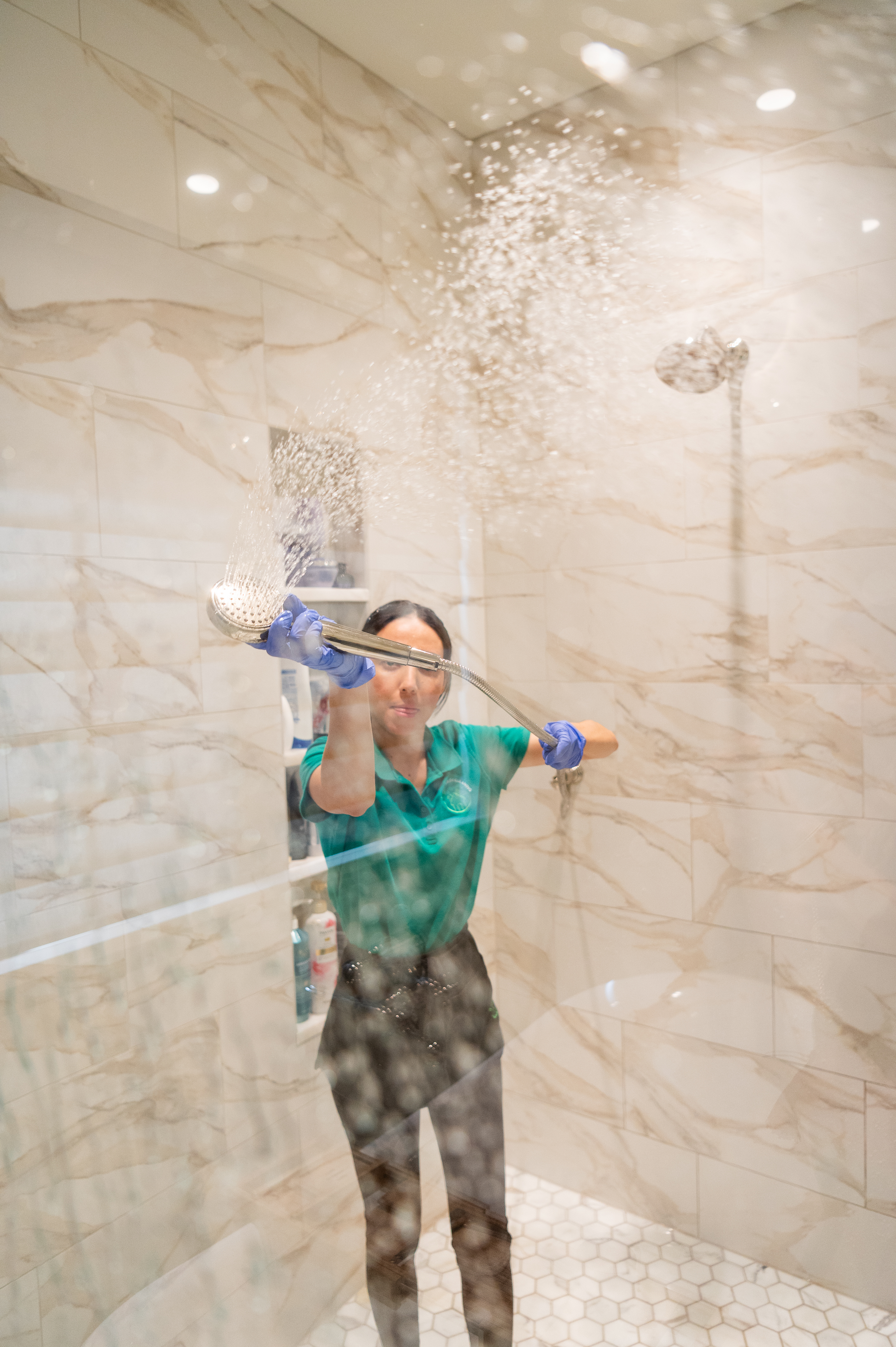 A professional cleaner wearing gloves and a green uniform thoroughly rinses a glass shower door using a handheld showerhead. The water spray highlights the cleanliness of the glass, demonstrating effective shower door maintenance.