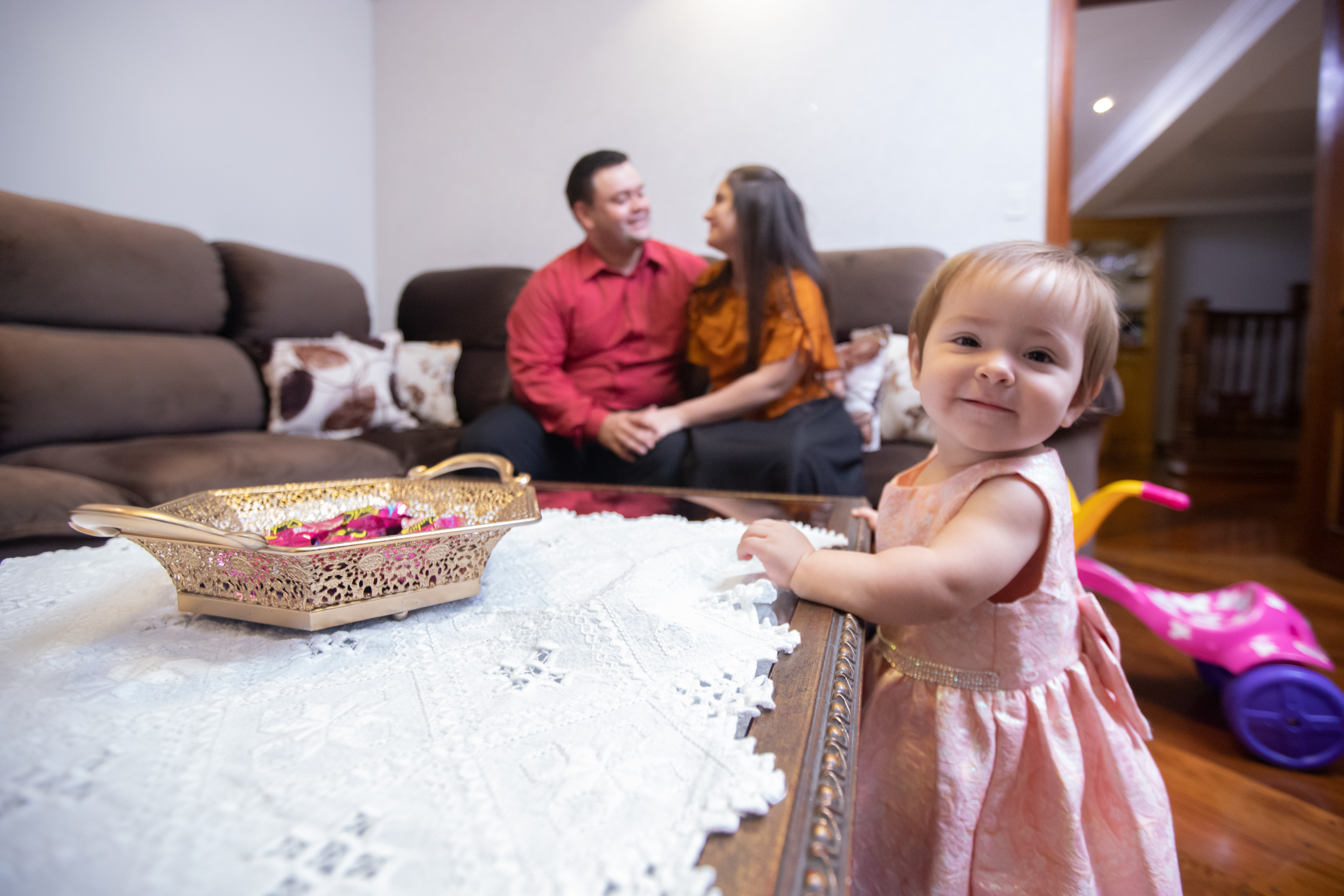 A happy family spending quality time together in a clean and organized living room. A baby in a pink dress smiles in the foreground, while the parents enjoy a relaxed moment on the couch. A tidy and welcoming home enhances family bonding.