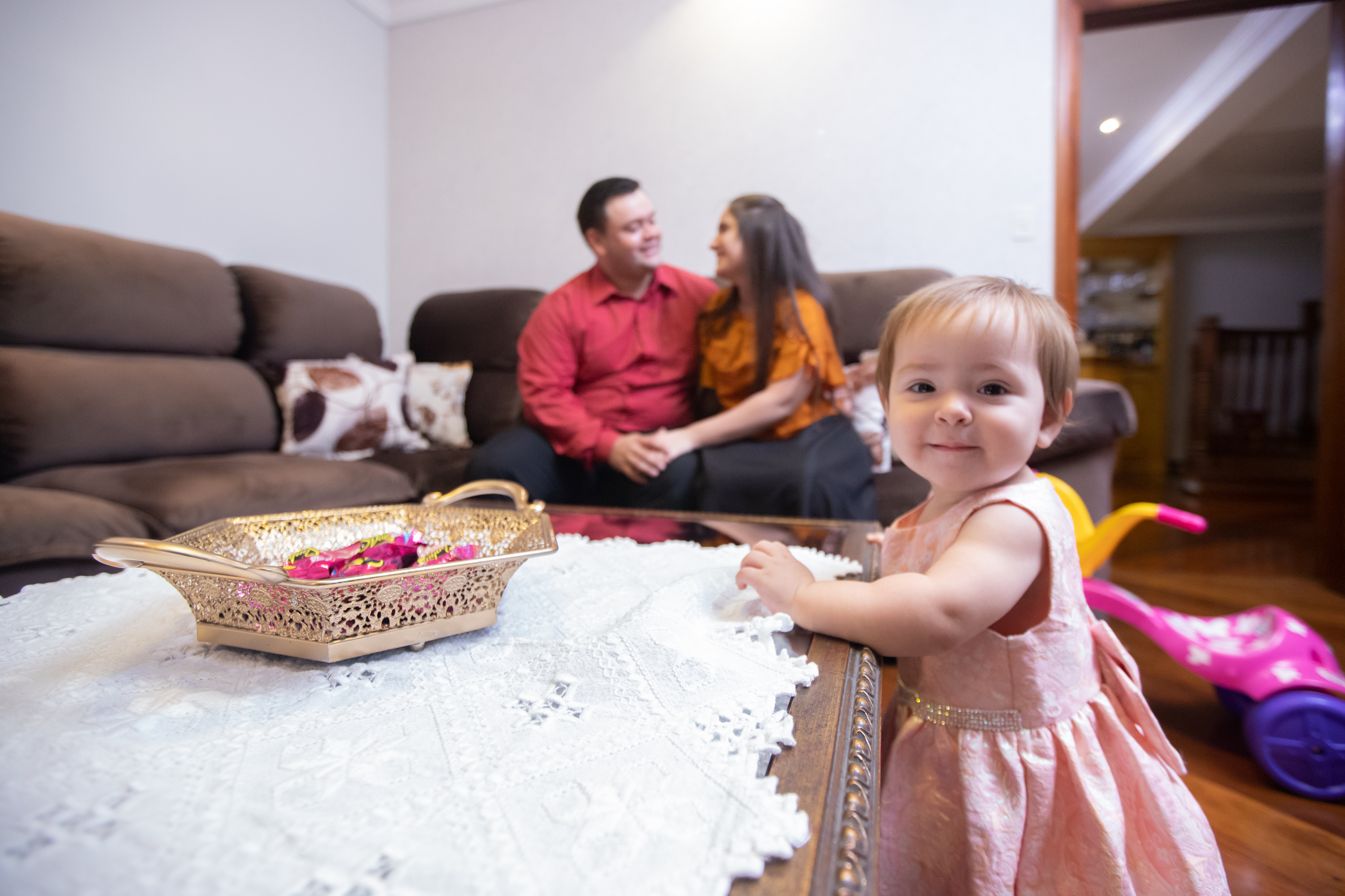A smiling toddler in a beautiful dress stands near a coffee table, possibly the culprit of a small milk spill. In the background, parents sit on a cozy couch, unaware of the potential mess. A warm, family-friendly scene that highlights the importance of quick milk spill cleanup to keep a home fresh and stain-free.