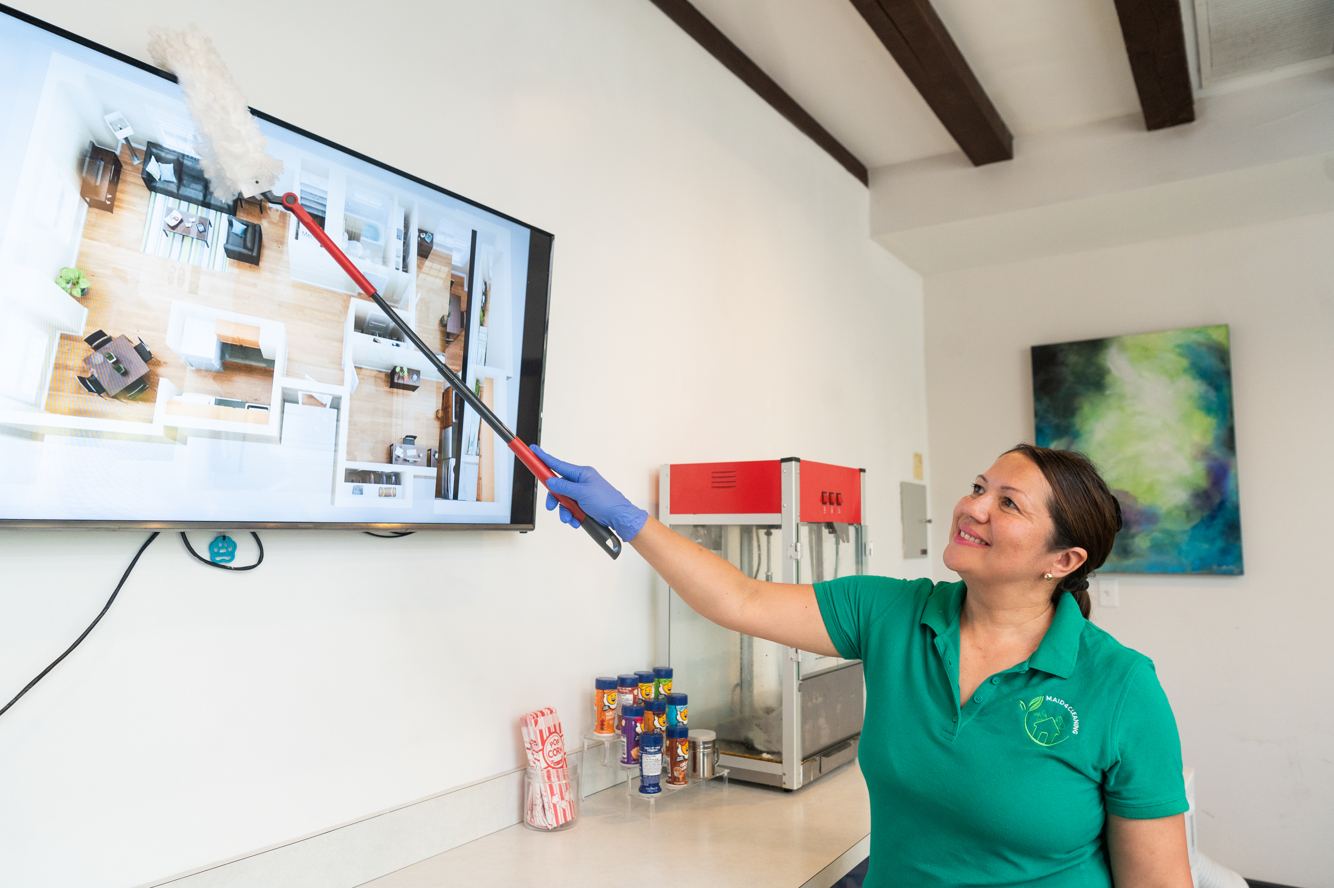 A professional cleaner wearing a green Maid 4 Cleaning uniform uses a microfiber duster with an extension handle to gently clean a mounted flat-screen TV. This demonstrates the importance of safe and effective TV screen cleaning, as outlined in our blog post on maintaining a streak-free and germ-free viewing experience for the Big Game.