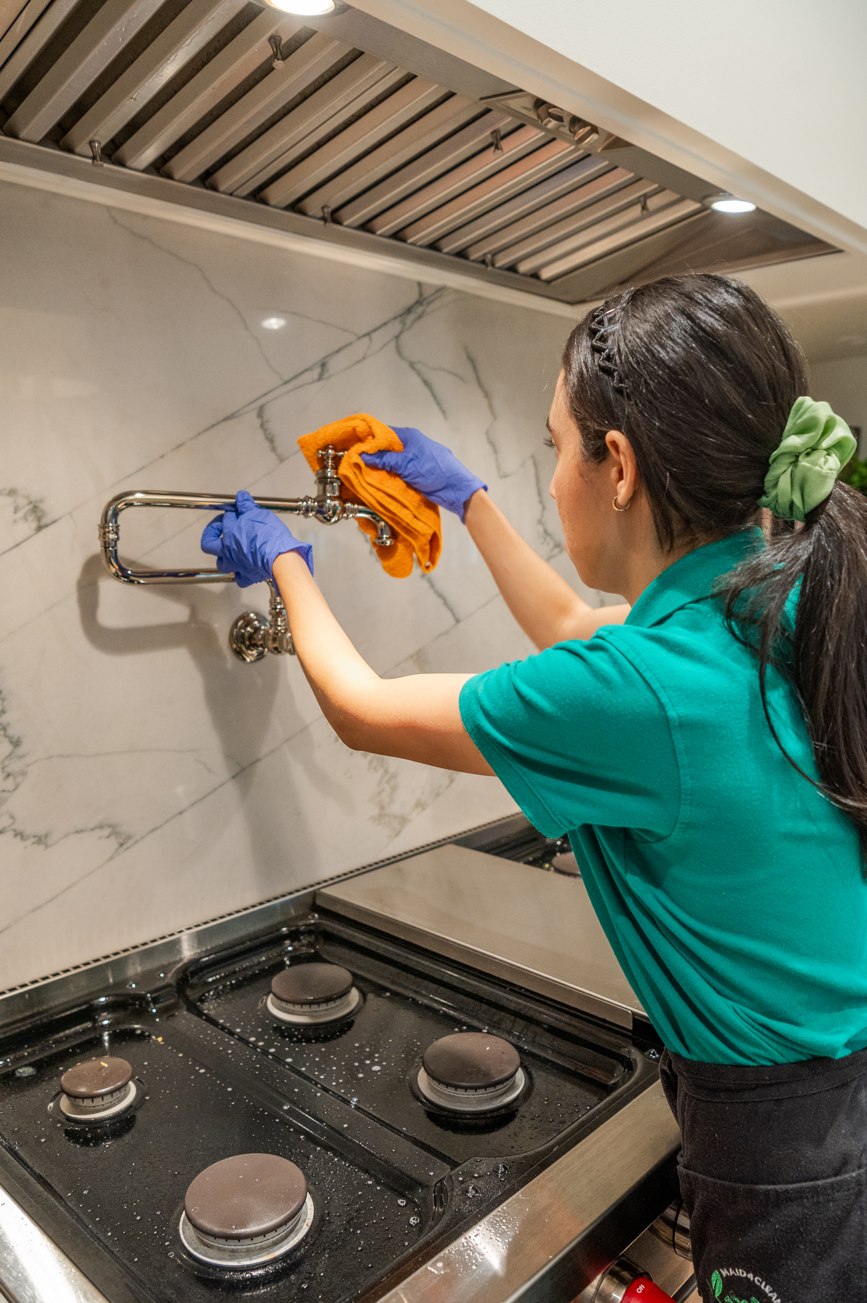 A professional cleaner wearing gloves and a green uniform wipes down a stainless-steel pot filler above a stove, ensuring a spotless and grease-free kitchen surface.