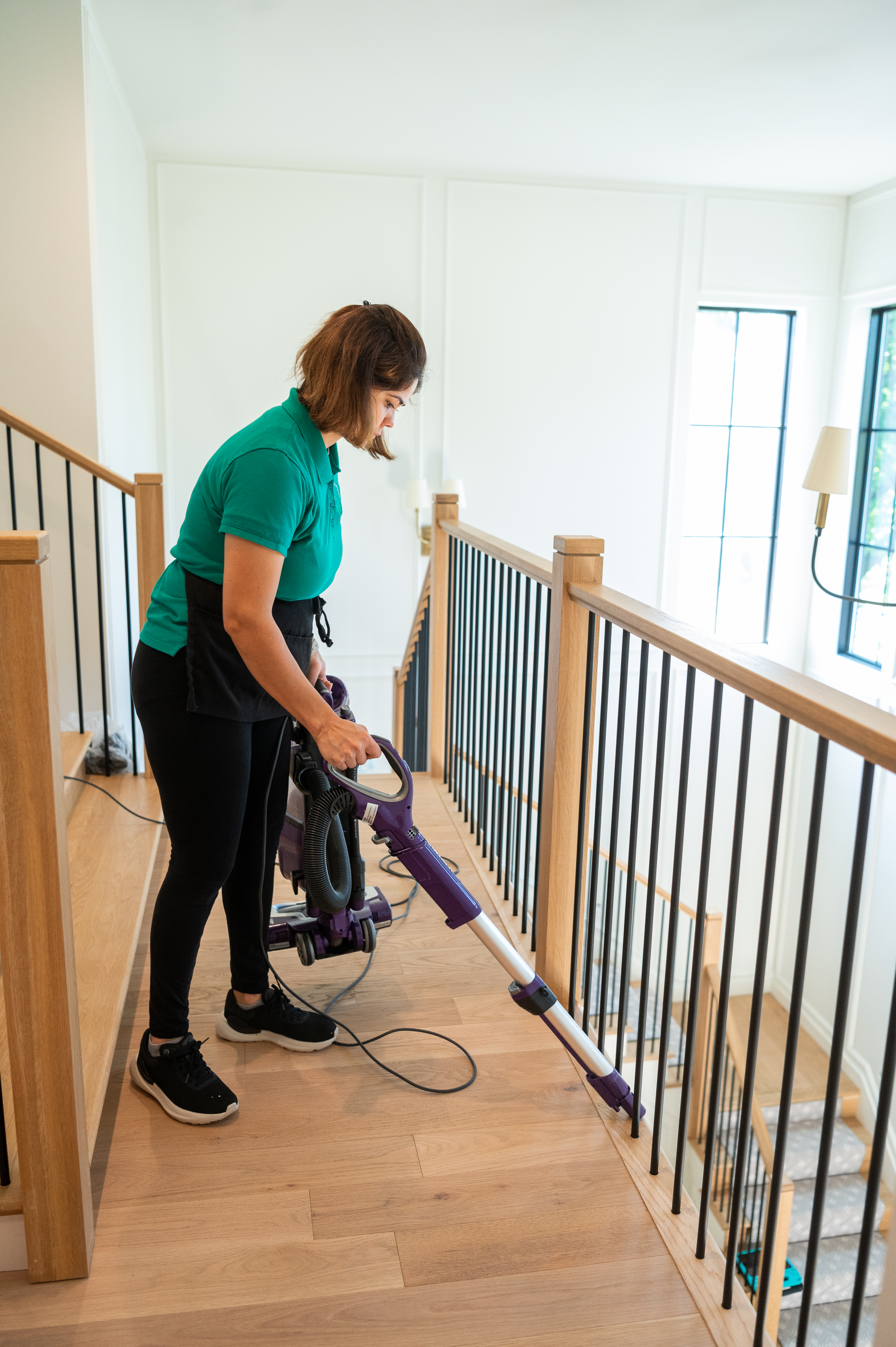 A professional cleaner from Maid 4 Cleaning using a vacuum with an attachment to clean dust and debris from a hardwood floor near a staircase, highlighting the benefits of vacuuming over sweeping for a thorough clean.