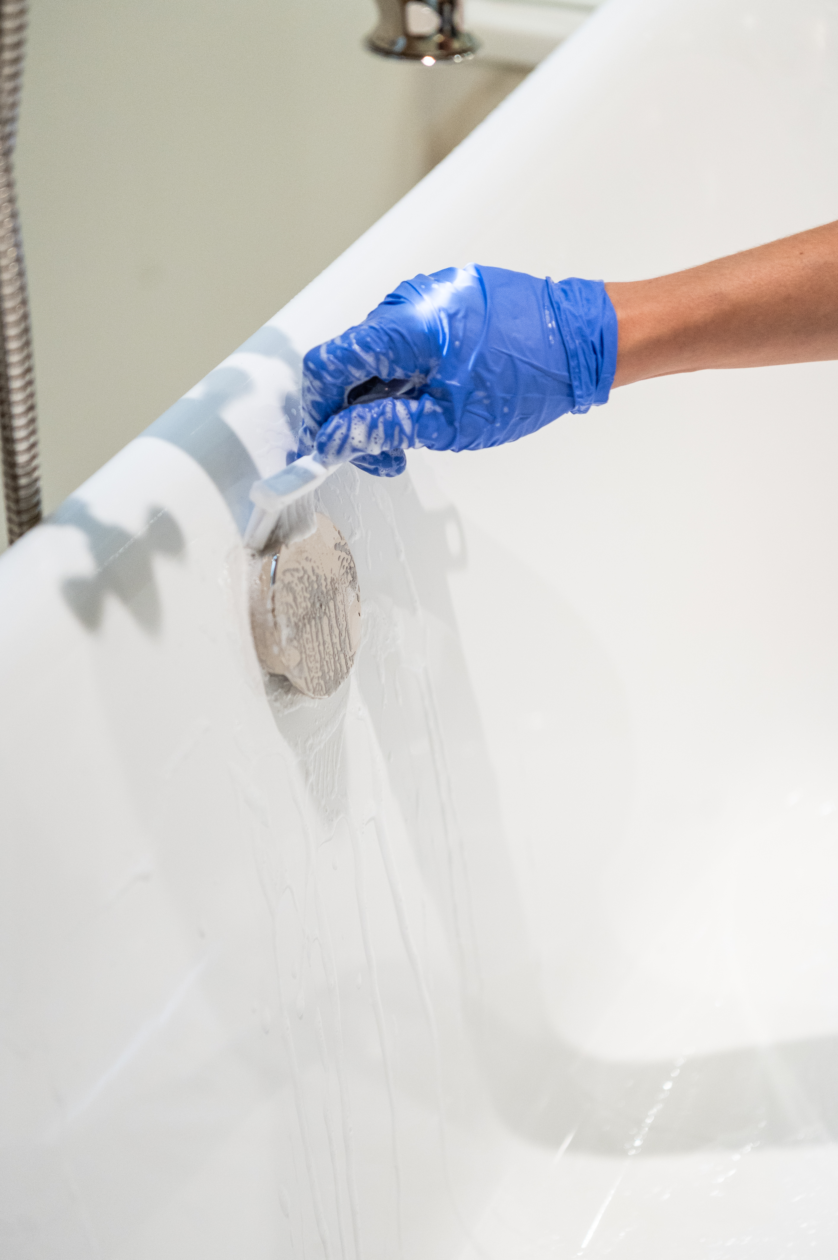 A person wearing blue rubber gloves scrubs rust off a bathtub drain using a small brush. This cleaning process helps remove stubborn rust stains and restore the fixture's shine, an essential step in maintaining a rust-free bathroom as outlined in our cleaning guide.