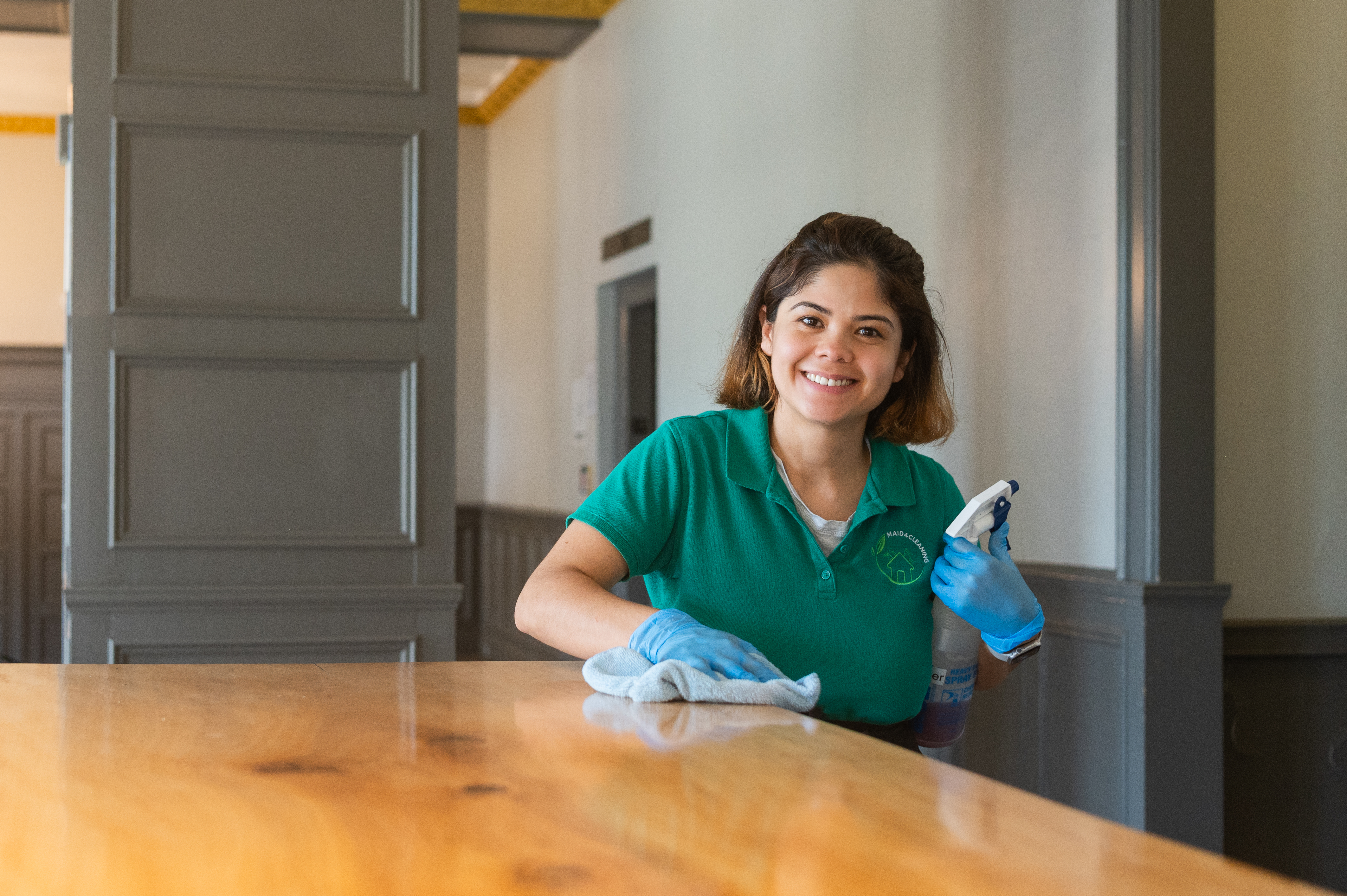 A professional cleaner wearing a green uniform and blue gloves smiles while wiping down a wooden surface with a microfiber cloth and a cleaning spray. She is working in a freshly cleaned, elegant interior, reflecting the thoroughness of post-construction cleaning services.