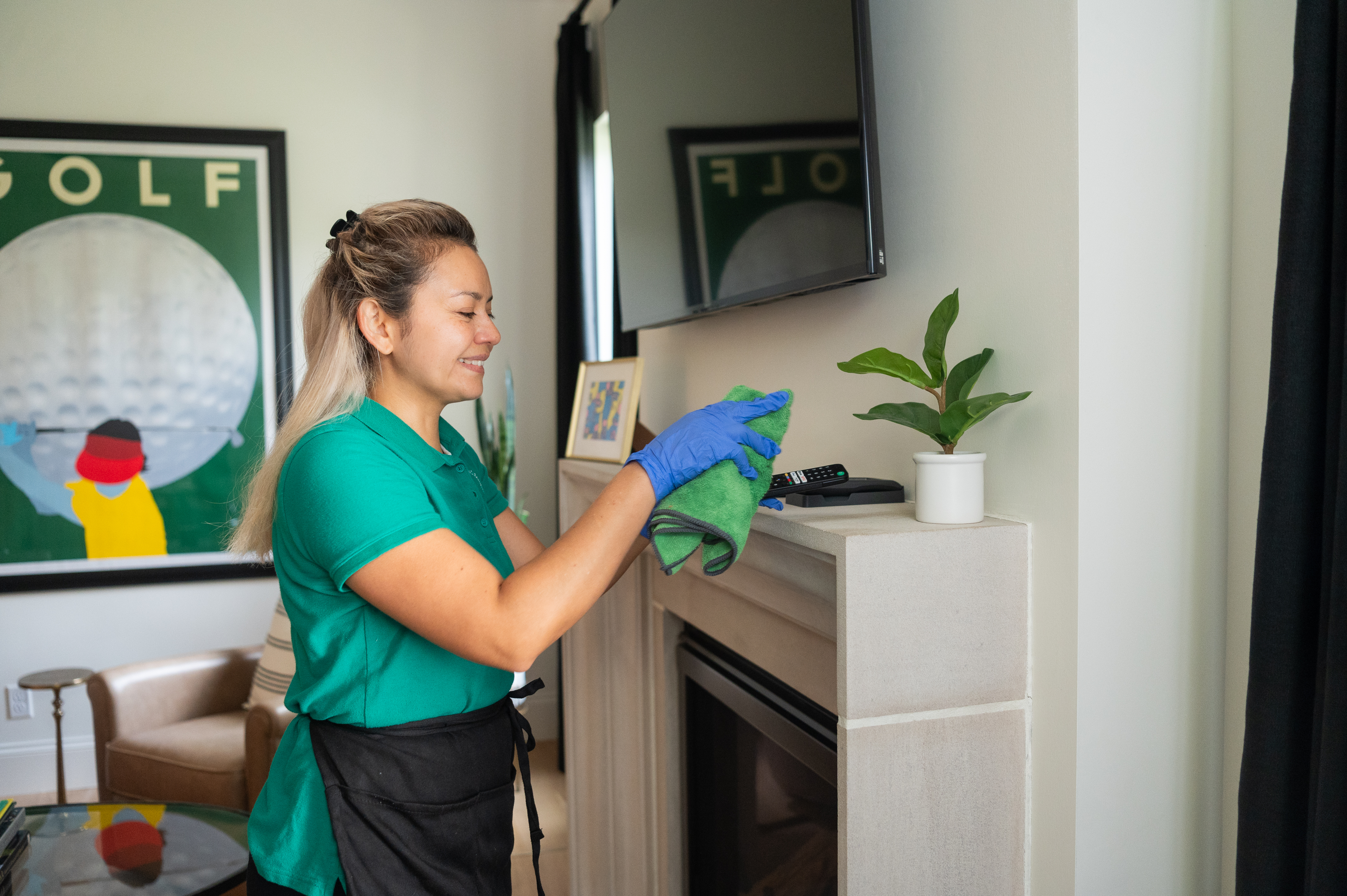 A professional cleaner wearing a green polo and blue gloves wipes down a fireplace mantel with a microfiber cloth. A small houseplant sits nearby, enhancing the clean and fresh home environment.