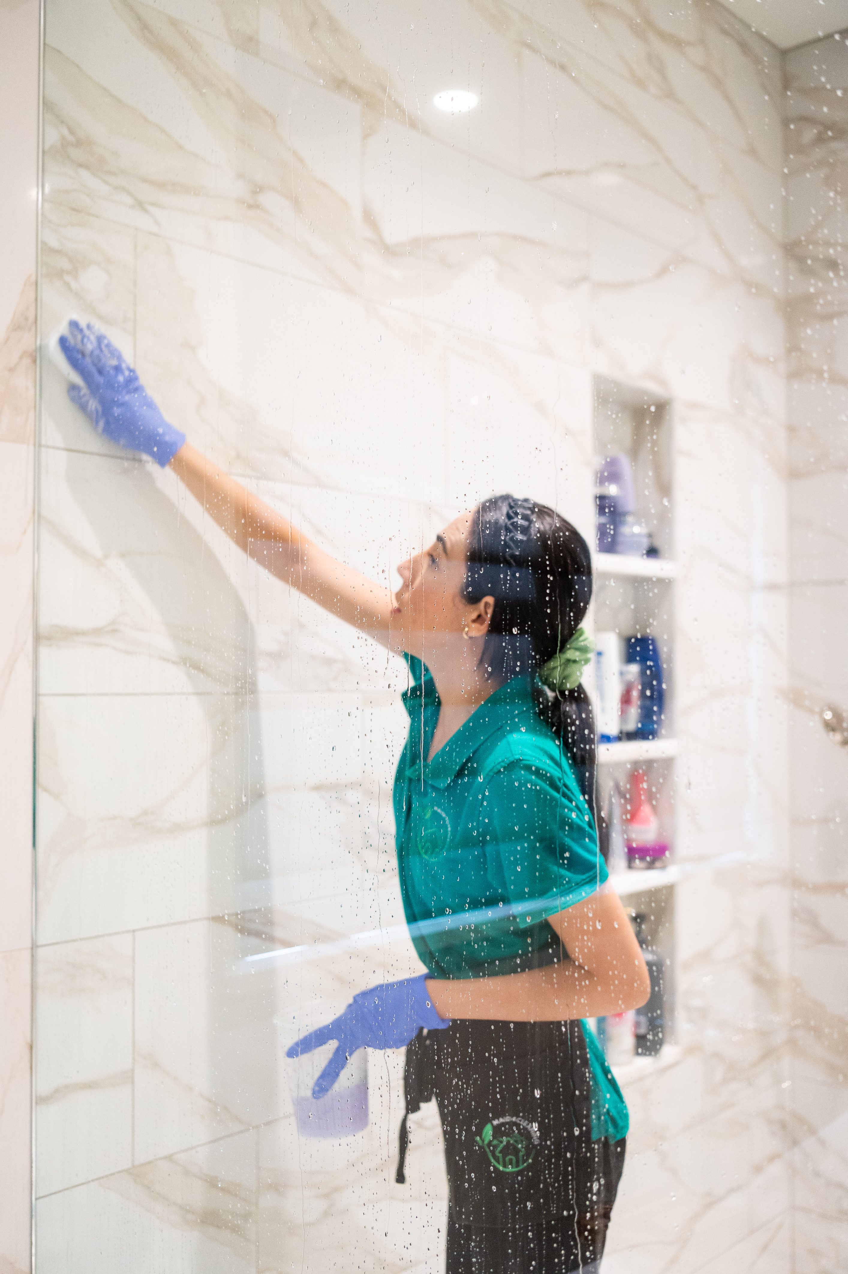 A professional cleaner from Maid 4 Cleaning wearing gloves and a green uniform carefully wiping down a marble shower wall to maintain its pristine appearance. The shower features elegant marble tiles, and cleaning supplies are neatly organized in the background.
