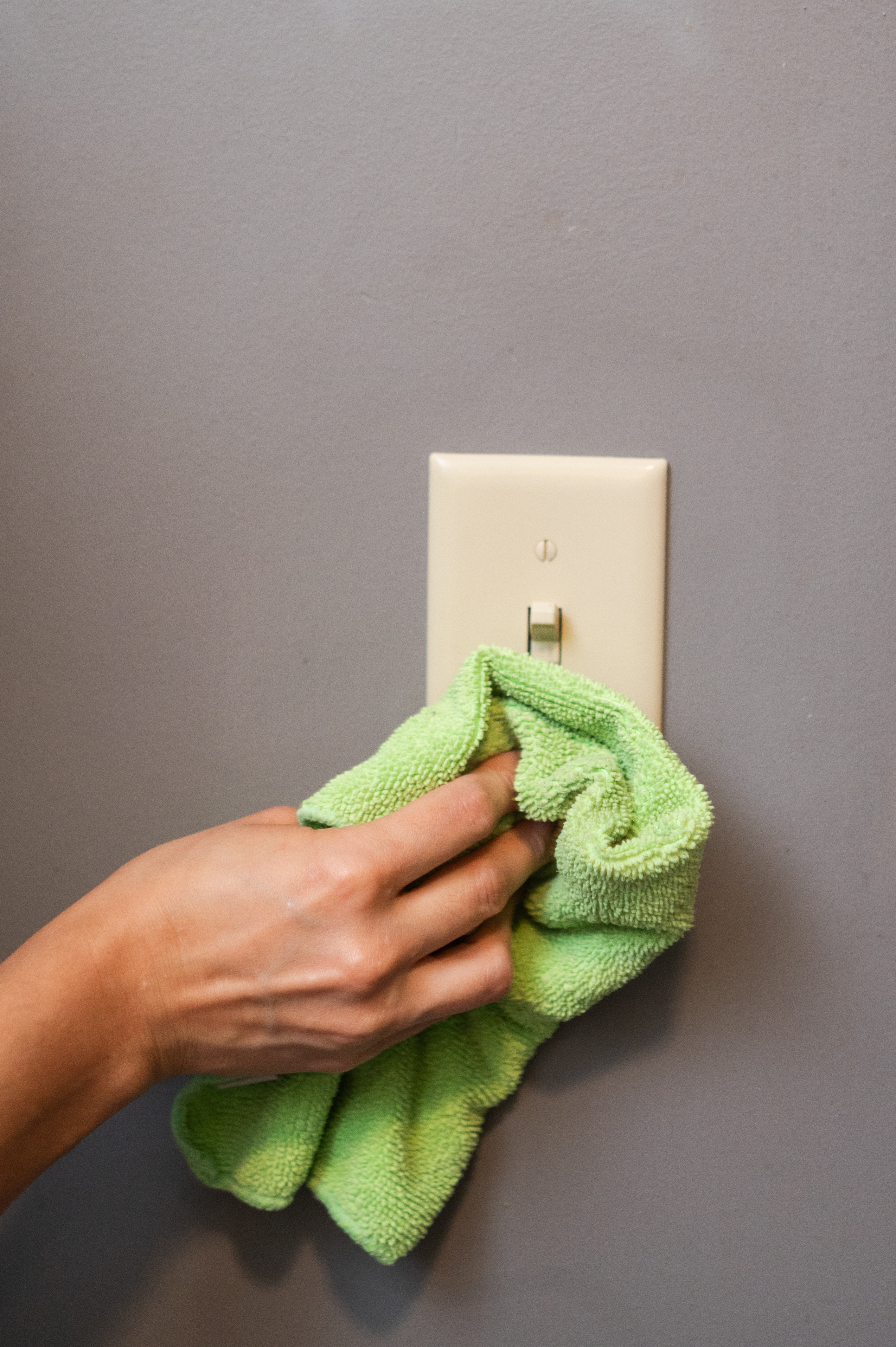 A close-up of a hand using a green microfiber cloth to clean a light switch, removing dirt and germs. This quick cleaning task, featured in the blog post, helps maintain a hygienic home and can be done in under 30 seconds.