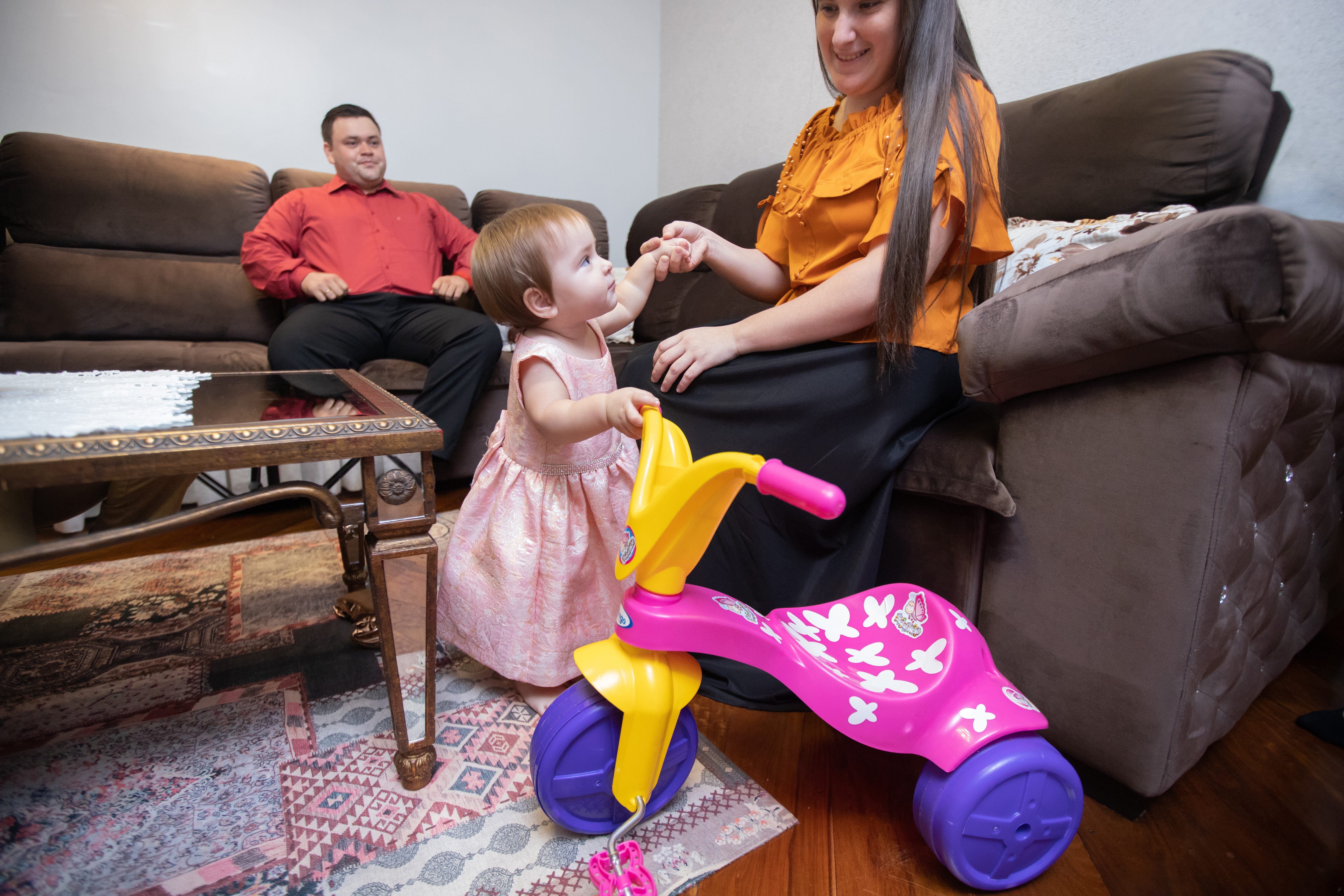 A toddler in a pink dress holds onto a colorful toy tricycle while interacting with her mother in a cozy living room, illustrating the importance of organized toy storage in shared family spaces.