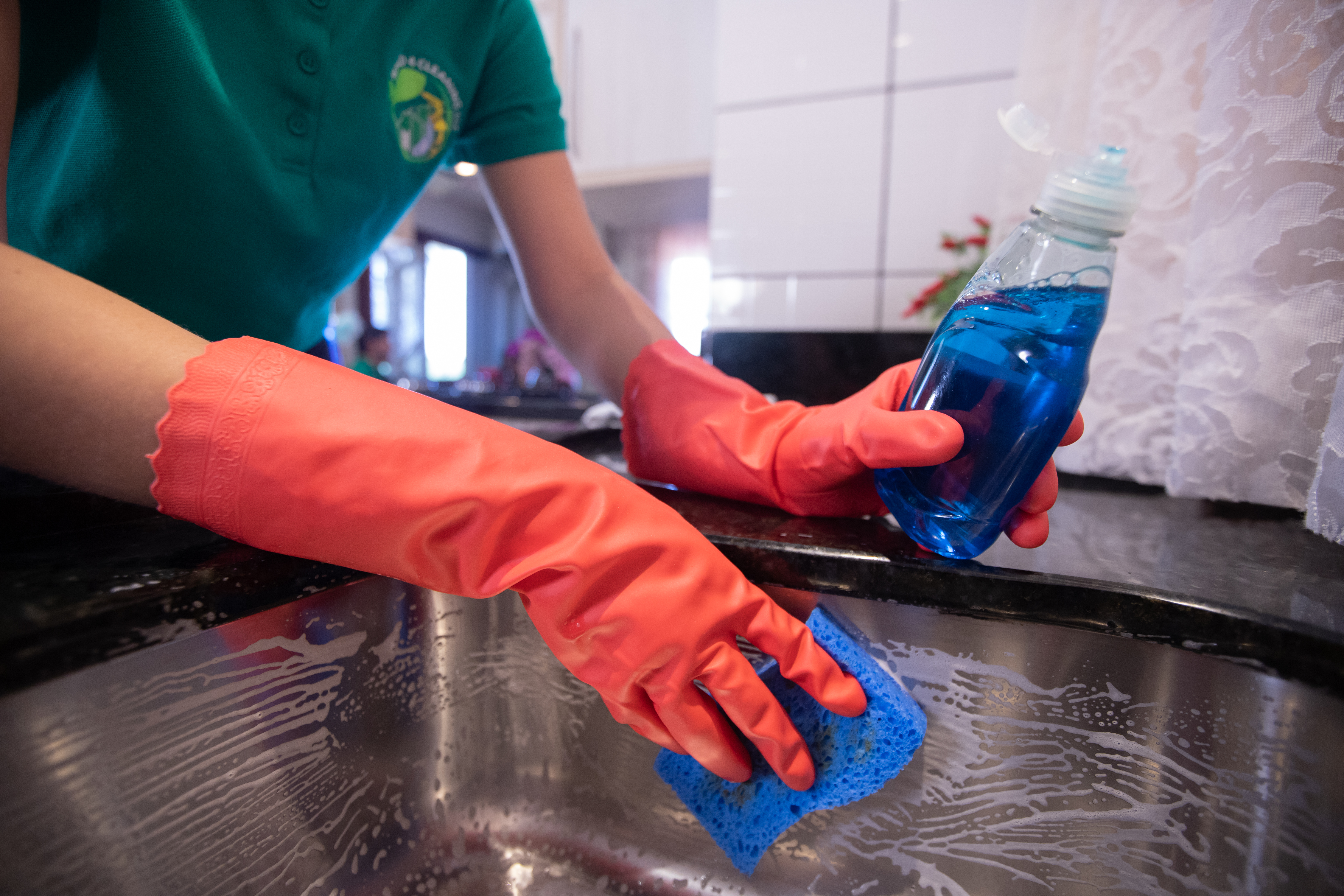 A professional cleaner wearing red rubber gloves scrubs a stainless steel sink with a blue sponge and dish soap, ensuring a fresh and odor-free kitchen.