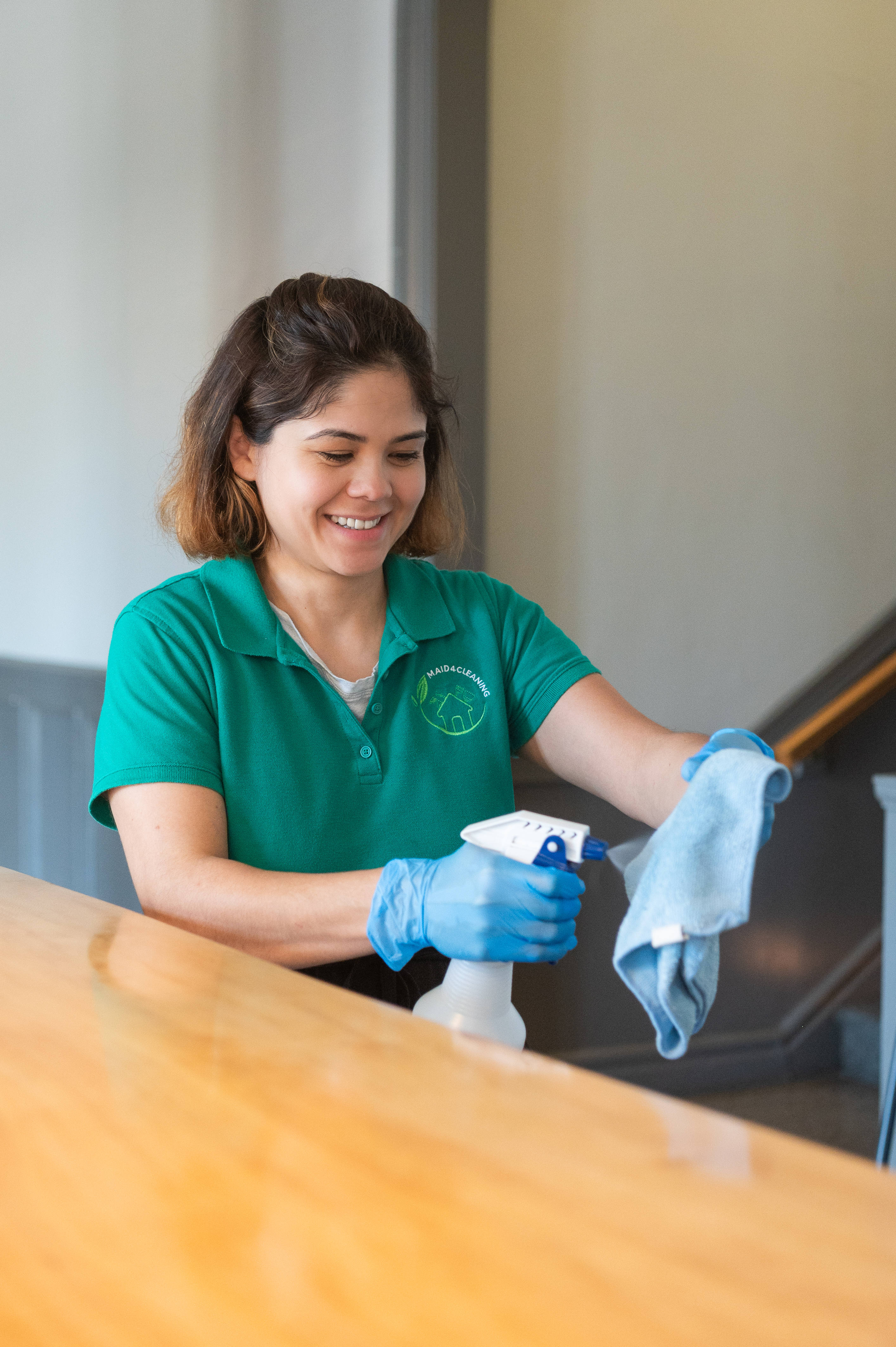 A professional cleaner from Maid 4 Cleaning in a green uniform smiles while wiping down a wooden surface with a microfiber cloth and cleaning spray, ensuring a spotless and tidy home.