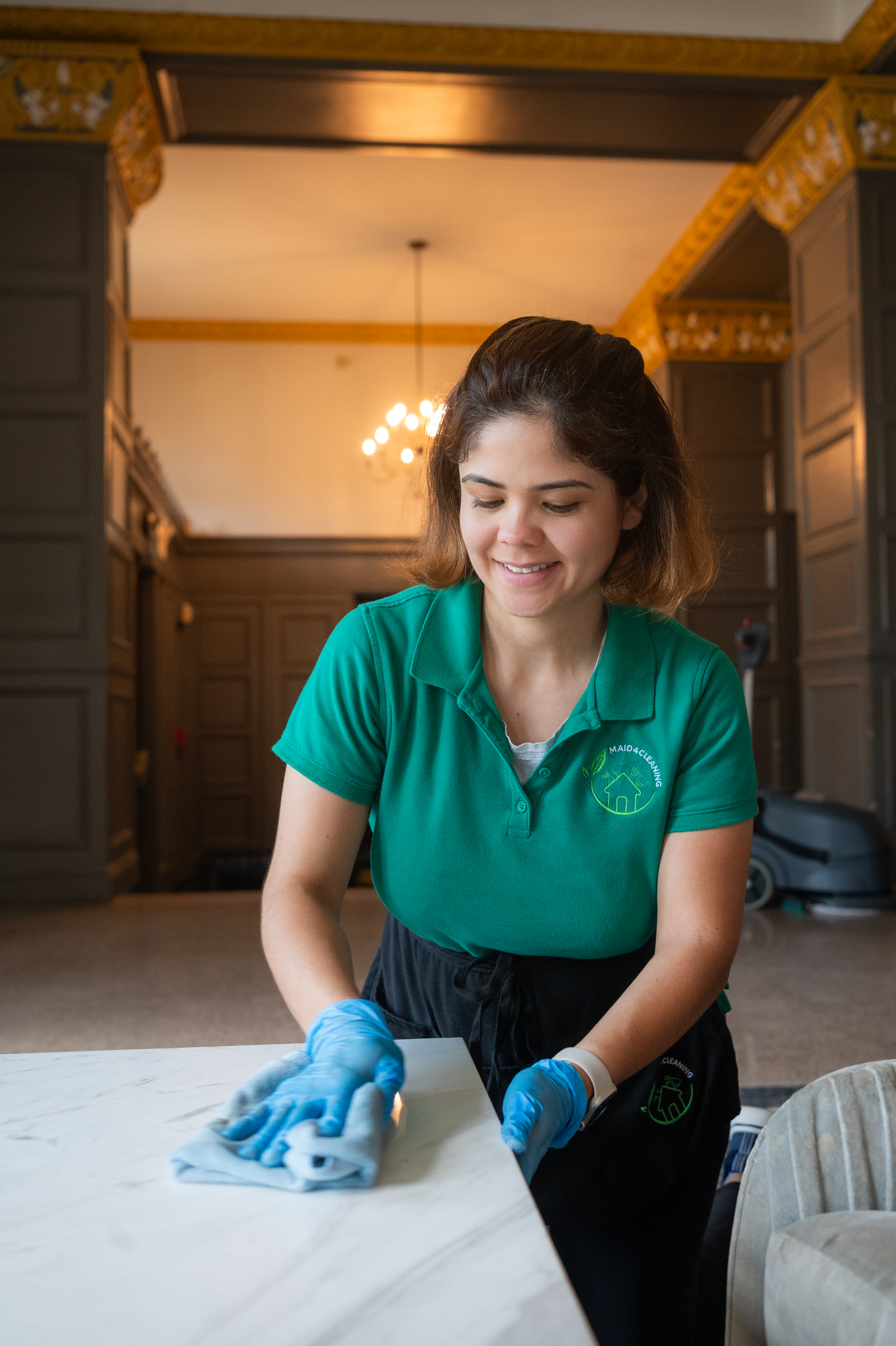 A professional cleaner from Maid 4 Cleaning wearing a green uniform and blue gloves, smiling while wiping down a marble countertop in a beautifully maintained space. Start the new year with a sparkling clean home!